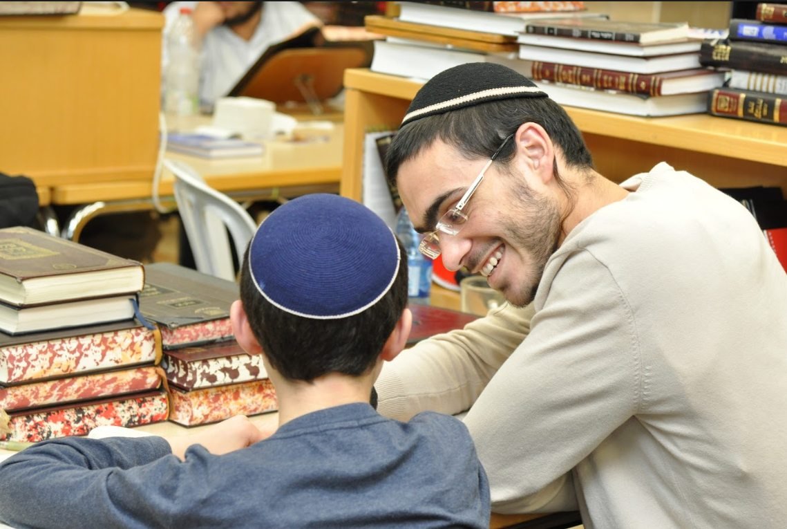 חונך מחייך בחום אל ילד שיושב לידו ליד שולחן עם ספרים